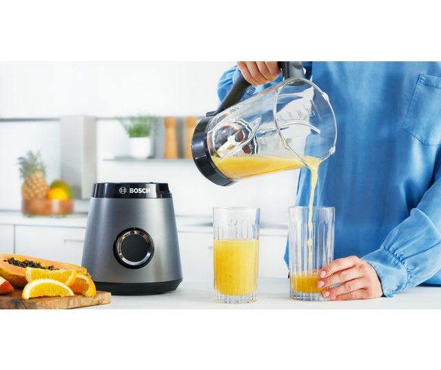 Bosch MMB6172S blender in gebruik, geel sap wordt uit glazen kan in glas gegoten op keukenblad